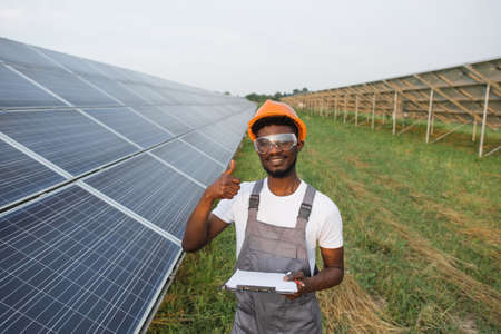 Portrait of african american man in safety glasses and orange helmet holding clipboard while standing on field with solar panels showing thumb up. Concept of eco energy and service workの写真素材
