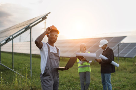 African american technician in overalls looking at camera while standing at solar station with...