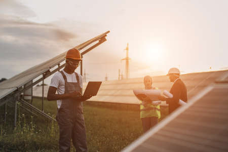 African american technician in uniform standing on field with solar panels and typing on laptop while muslim woman and indian man working with blueprints behind. Alternative energy concept.の写真素材