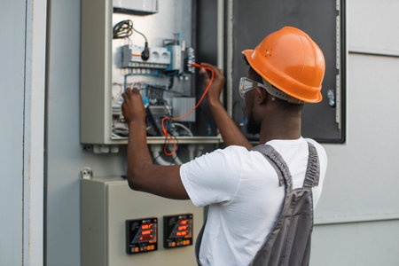 African american repairman in orange helmet and safety glasses checking cables in power transformer...