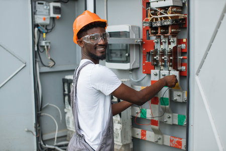 African american man in grey overalls, safety helmet and glasses smiling and looking at camera while standing near switchgear at solar station. Production of alternative energy.の写真素材