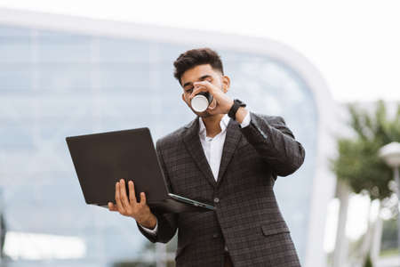 Focused young successful Arab businessman in formal wear, standing outside modern building and surfing internet on laptop while drinking coffeeの写真素材