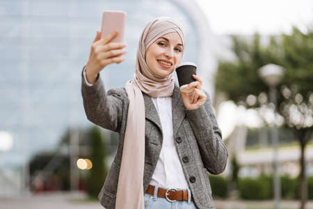 Young Muslim businesslady economist in formal wear, holding a cup of coffee while speaking on a smartphone. Young thoughtful Arab woman standing outside modern building and drinking take away coffeeの写真素材
