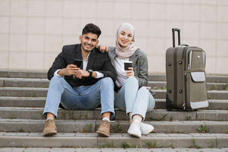 Two multiracial business people sitting at stairs with travel bag and looking at camera. Diverse colleagues ready for the trip, sitting with phones and coffee on strairs outsideの写真素材
