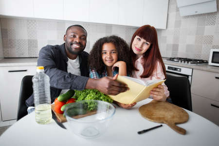 Book about healthy food. Happy adorable multiracial family of three looking at the camera with pleasure smiles while preparing fresh salad at the kitchen. Stock photoの写真素材