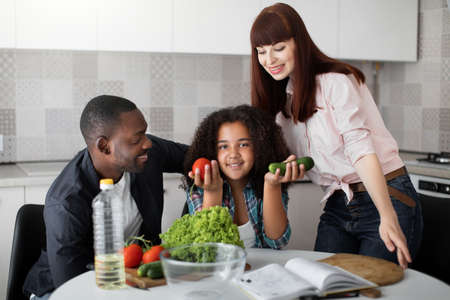 Lovely caucasian mother and black father teaching their teenage daughter to prepare salad for dinner lunch. Cute mixed race girl holds fresh organic bio vegetables, tomato and cucumberの写真素材