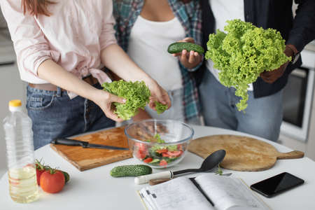 Cropped view of the teen girl preparing vegetables for the future salad on chopping board while standing at the light kitchen with her parents. Stock photoの写真素材