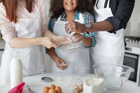 Cropped view of the happy father, his wife and teen girl baking cookies, pie or preparing pancakes for breakfast at home. Caring young family cooking in modern kitchen togetherの写真素材