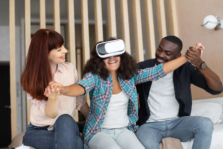 Pretty emotional teenage girl outstretching hands while trying VR headset and sitting at the bedroom, spending time with her multiracial mother and father. Stock photoの写真素材