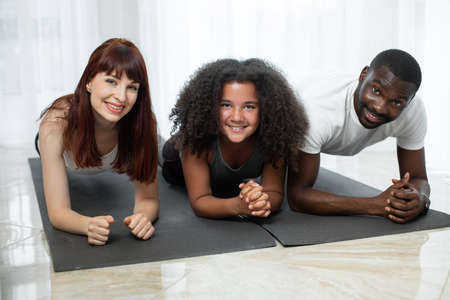 At plank. Handsome multiracial parents doing plank with their daughter at the yoga mat. Parents including child in activity. Family quarantine, domestic life in self-isolation conceptの写真素材