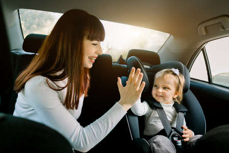Pretty young woman mother takes care about her little daughter in a car safety seat, sitting together, giving high five each other and having fun. Safety road, happy family trip, baby safety.の写真素材