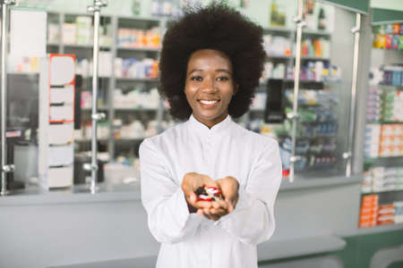 Young smiling African-american female pharmacist, holding a handful of medicine pills and colorful tablets in the hands, offering to camera. Drug store and drug quality concept. Focus on face.の写真素材