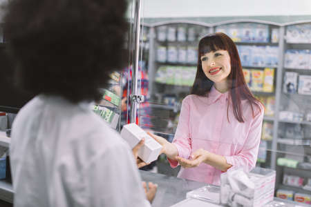 Back view of young african female pharmacist selling medicines to young beautiful Caucasian woman patient. Drug dispensing at modern pharmacy.の写真素材