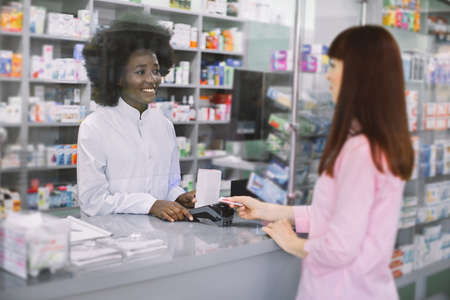 Pretty young Caucasian woman paying for mediciens with credit card in pharmacy. Smiling attractive African woman pharmacist dispensing drugs for young woman at pharmacy.の写真素材