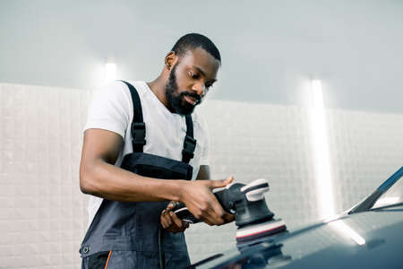 Car detailing service. Handsome bearded African American worker in white t-shirt and gray overalls, holding orbital polisher in repair shop while polishing the car. Selective focusの写真素材