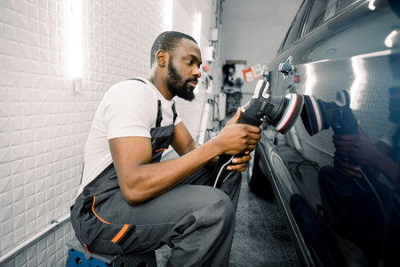 Auto detailing service, polishing of the car. Side view of young African American man worker n t-shirt and overalls, polishing blue car door with orbital polisher.の写真素材