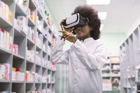 Bottom angle view of young pretty African American female clinical pharmacist or doctor, working in modern hospital drugstore in virtual reality goggles.の写真素材