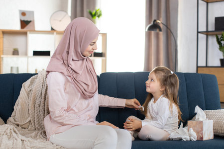 Happy loving Arabian family. Muslim mother in hijab is combing her little daughters hair, sitting on the sofa in cozy living room. Happy mother doing her daughters hairstyle and talkingの写真素材