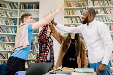 Multiethnic group of four satisfied pleasant successful students studying together in the university library, having good results on exam and giving each other high five. Education concept.の写真素材
