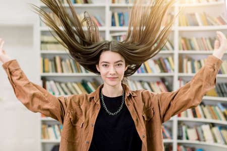 Funny portrait of pretty brunette young Caucasian girl, student, in brown casual shirt, posing on the book shelves interior of library, tossing her long straight hair up. Happy book and education.の写真素材