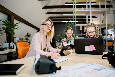 Group of young creative designers, business people, gathered together to discuss idea, develop startup. Two pretty girls and handsome guy working at the office table using electronics gadgets.の写真素材