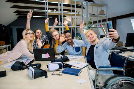 Handsome joyful man in wheelchair sitting at desk with his colleagues, and making funny selfie photo on smartphone. Group of multiethnic hipster coworkers posing for photo and waving.の写真素材