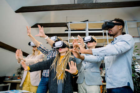 Office team of multiethnic colleagues wearing vr headset having fun together, taking part in virtual conference or webinar, touching virtual screen and gesturing. People working with new technologies.の写真素材