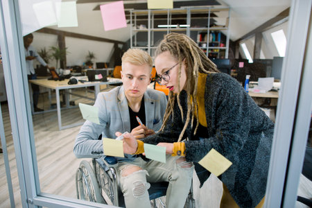 Two happy creative concentrated businesspeople, disabled man in wheelchair and pretty blond girl...