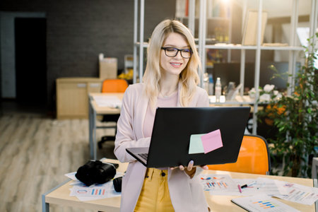 Concentrated young beautiful blond Caucasian businesswoman working on laptop in bright modern office. Pretty woman in glasses working on laptop in the office to finish the project.の写真素材