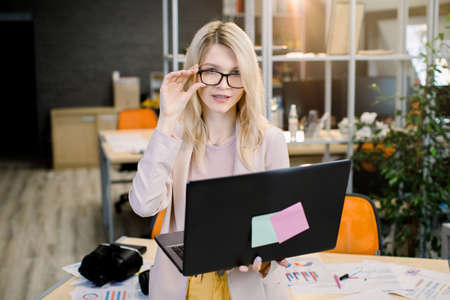 Beautiful young blond business lady in eyeglasses, wearing smart casual clothes, is looking at camera and smiling while working in modern creative office, using laptop.の写真素材