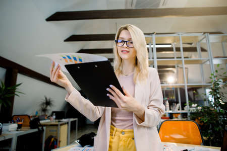 Attractive young business lady in trendy glasses and casual wear, holding folder with financial charts, posing in office preparing for meeting with colleagues. Indoor portrait of businesswomanの写真素材