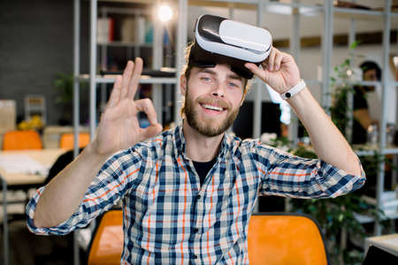 Business, people, 3d vision and success concept. Happy smiling bearded businessman in casual checkered shirt, having fun while testing new 3d vr glasses and showing ok sign over office backgroundの写真素材