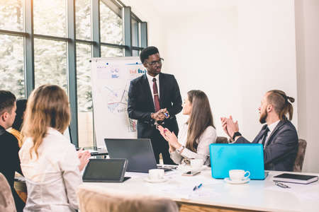 Business team talking to african-american applicant at work interview in office. young Afro American Man standing and makes a report near whiteboardの写真素材