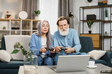 Joyful mature married couple with wireless joysticks in hands playing video games while sitting on...