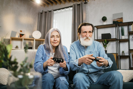 Positive pleasant old wife and her bearded husband holding console playing games while sitting...