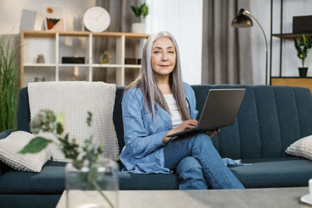 Pleasant attractive grey-haired woman with long hair sitting on couch with laptop on knees. Happy female pensioner enjoying remote work from home. Concept of retirement and free time.の写真素材