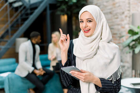 Pretty young Muslim business lady in white hijab standing in the modern loft office room, holding...