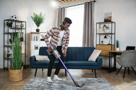 Smiling afro american man in wireless headphones cleaning carpet with handheld vacuum cleaner. Young guy enjoying housekeeping using modern devices.の写真素材