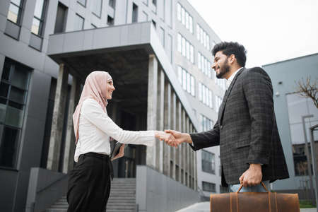 Side view of muslim business partners in formal clothes standing on street and shaking hands. Young woman and man having successful common project. Deal concept.の写真素材