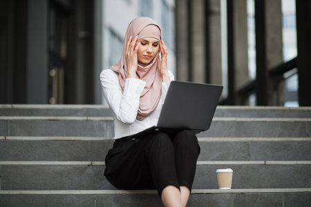 Arabian woman in hijab sitting on stairs with modern laptop on knees and touching her temples...