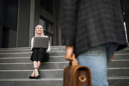 Smiling woman in hijab and formal wear sitting with modern laptop on knees. Close up of man with suitcase going up on stairs. Two partners coworking outdoors.の写真素材