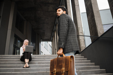 Handsome muslim man in formal wear coming up on stairs while woman in hijab sitting near and working...