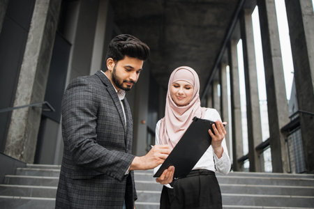Attractive woman in hijab holding clipboard while handsome hindu man signing contract. Two business...