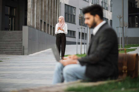 Blur foreground of muslim man in stylish wear sitting on bench and working on laptop. Pretty woman in hijab walking on background with smartphone on hands. Urban area.の写真素材