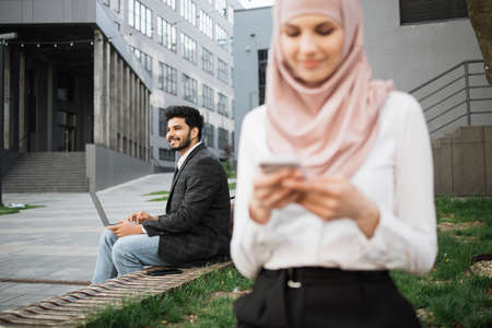 Blur foreground of muslim woman in hijab sitting on bench and using smartphone. Handsome man working on wireless laptop on background. Modern gadgets for remote work.の写真素材