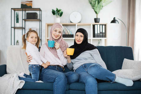 Three muslim female generations family portrait. Cute girl, islamic mom and grandma hold mugs of hot drinks. Mother, grandmother and granddaughter on couch at home, enjoying leisure time togetherの写真素材