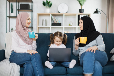 Old muslim granny young mom holding mugs of hot drinks with little girl having fun with laptop,...