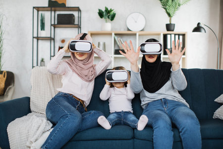Happy three generations of muslim women watching in modern augmented vr headset reality glasses In...