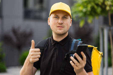 Close up portrait of delivery man in yellow cap with thermo backpack checking orders using smartphone while riding an electric scooter. Courier, delivery service concept. Horizontal shotの写真素材
