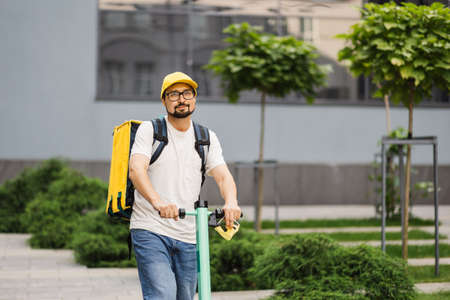 Food delivery electric scooter driver with yellow backpack behind back is on his way to deliver food. Courier on scooter delivering food. Quick shipping of goods to customersの写真素材